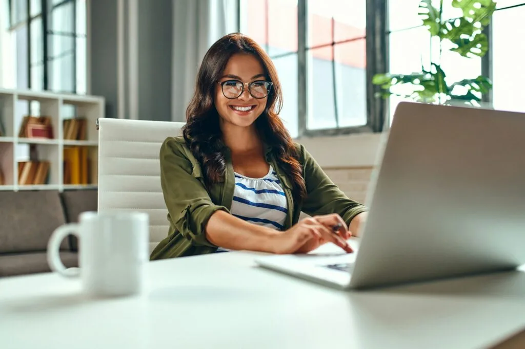 Smiling woman wearing glasses working on a laptop at a desk in a bright home office, with a coffee mug in the foreground and large windows and plants in the background, representing online loan application or digital financial management.